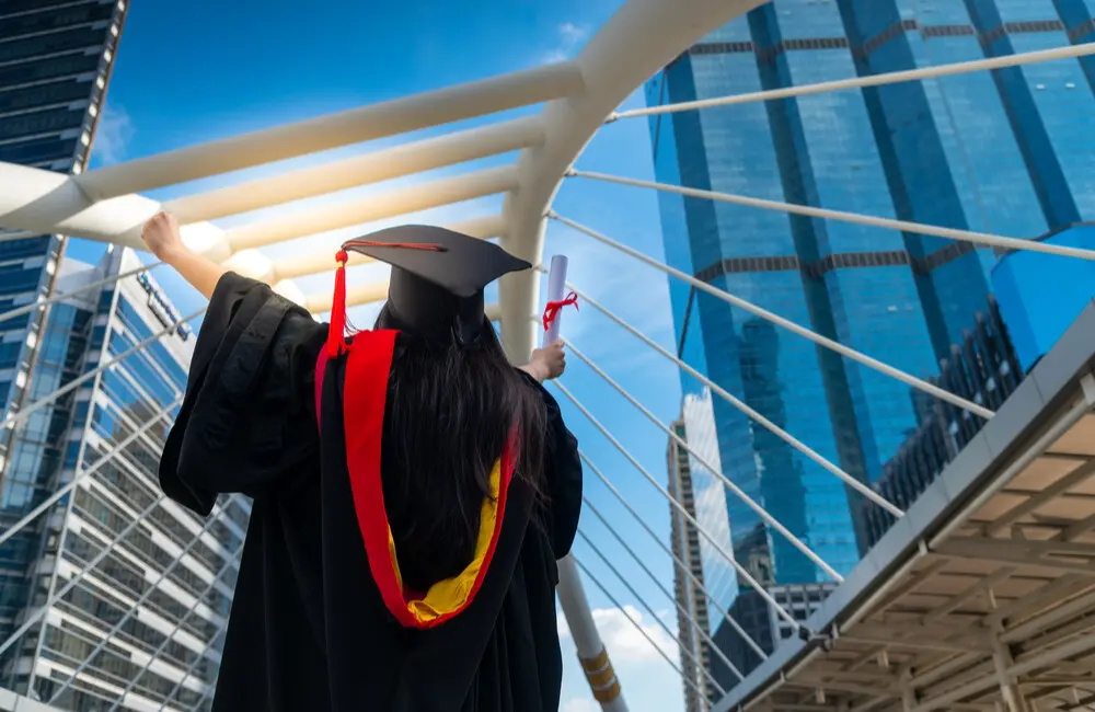A graduate wearing a cap and gown and holding a diploma raises her arms to the blue sky in the middle of a city block.