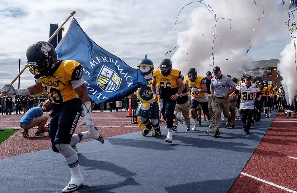 The Merrimack football team runs out onto the field carrying a Merrimack flag.