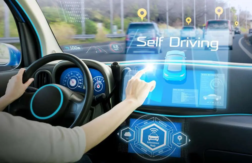 A person holds the wheel of a car with one hand and uses a touch screen on the console with the other to activate Self Driving mode.