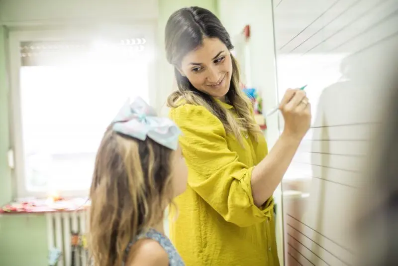 A teacher smiles at a student standing beside her as she writes on a whiteboard.