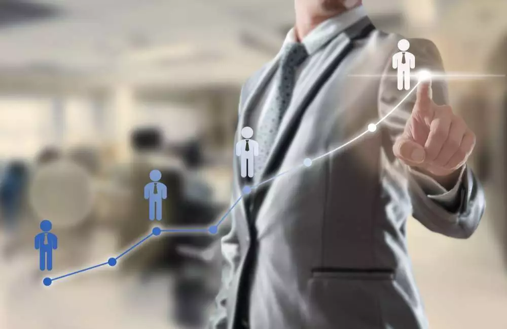 A man in a suit points to a transparent screen with a line graph of employee icons that transform from dark blue to white.
