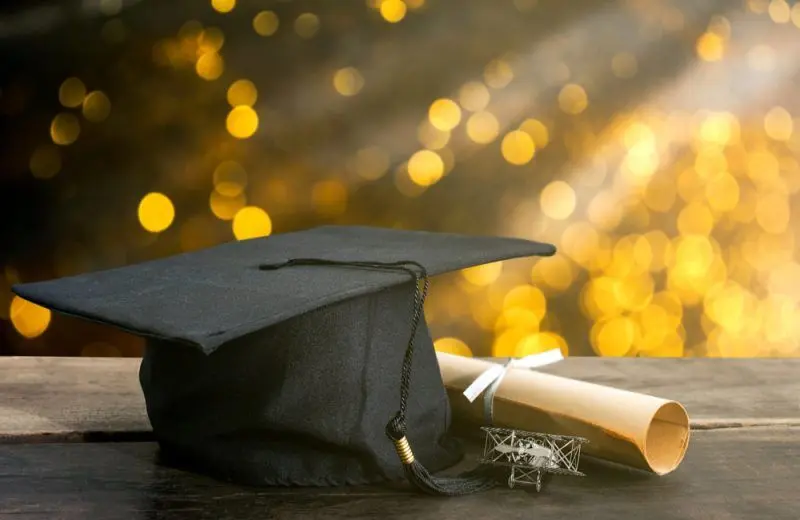 A graduation gap and diploma on a wooden table with warm yellow lights in the background.