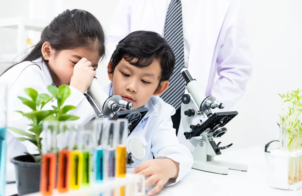 Two young students wear lab coats and look through a microscope.