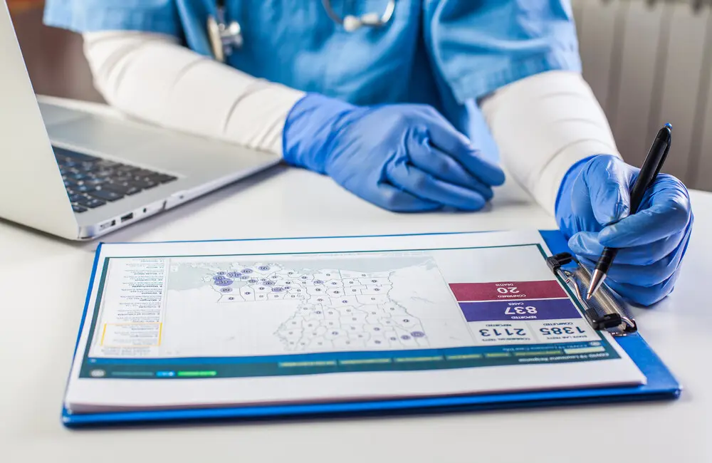 A healthcare professional wearing gloves and holding a pen looks a clipboard of data.