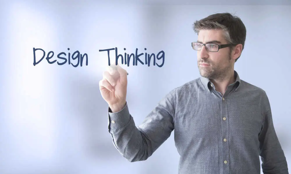 A man wearing glasses writes the words Design Thinking on a transparent screen.