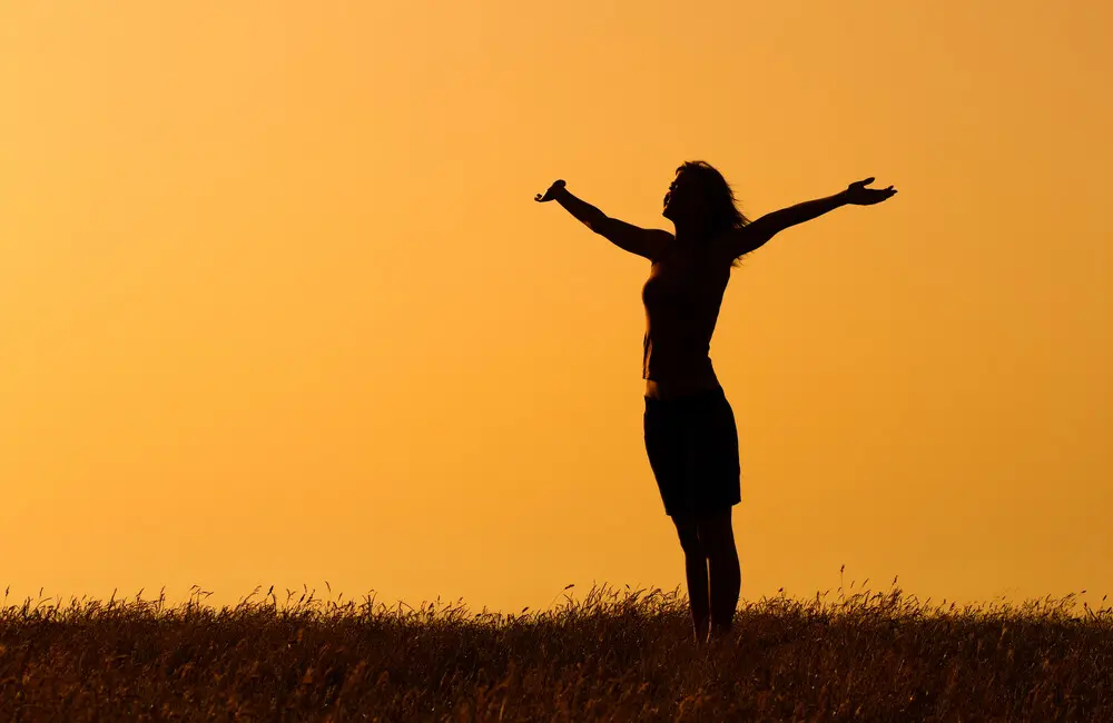 A woman in silhouette stands in a field of grass with her arms fully extended.