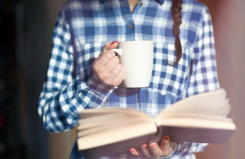 A closeup of one hand holding a coffee mug while the other hand holds an open book.