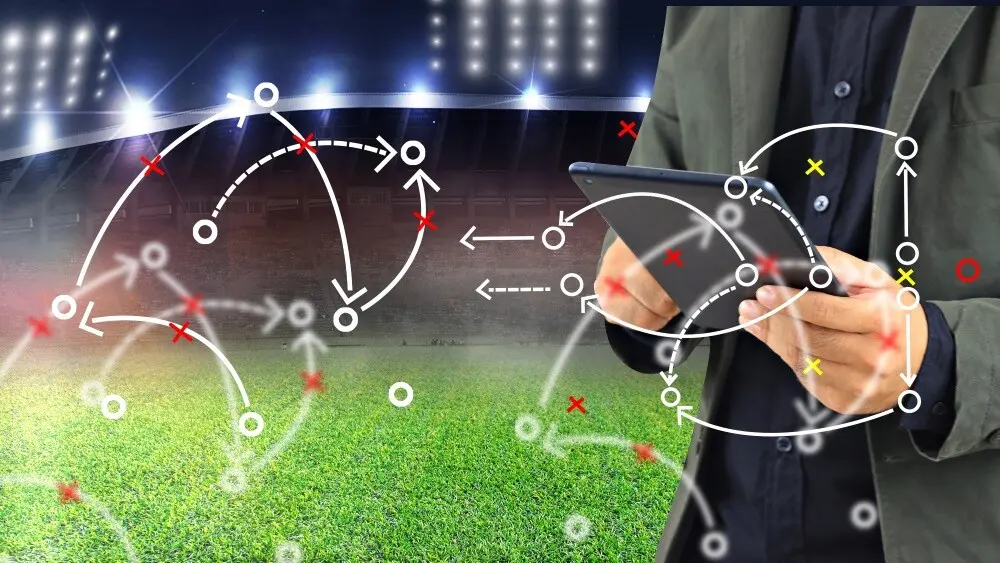 A closeup of hands holding a tablet with multiple football playbook notations in the foreground.