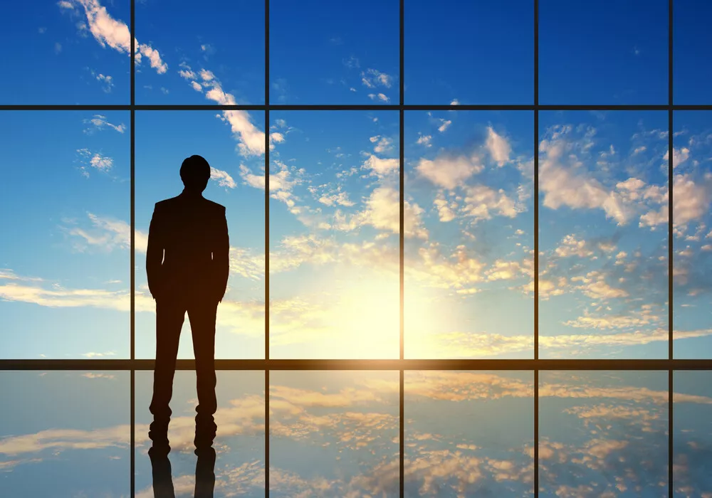 A man in a suit stands in silhouette looking out floor to ceiling windows at a blue sky.