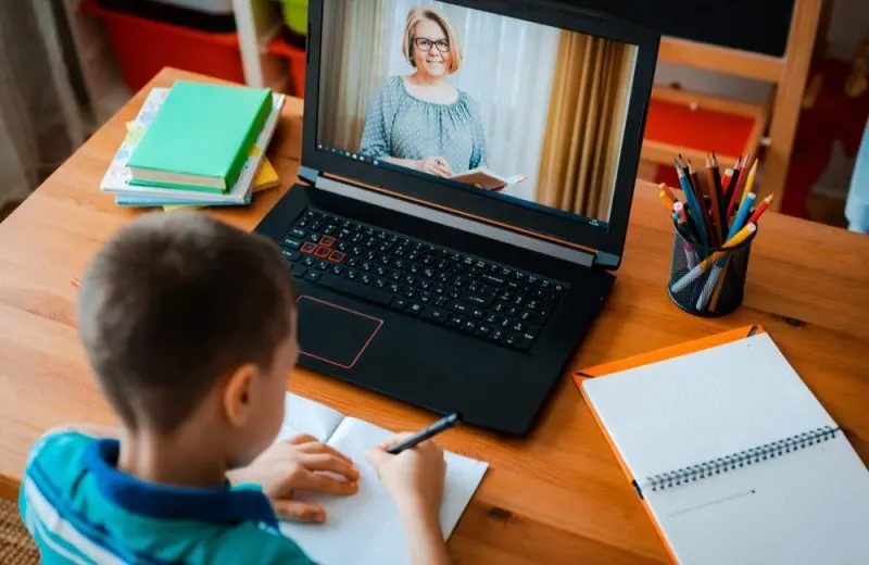 An aerial view of a young child writing in a notebook in front of a open laptop displaying a video call with his teacher.
