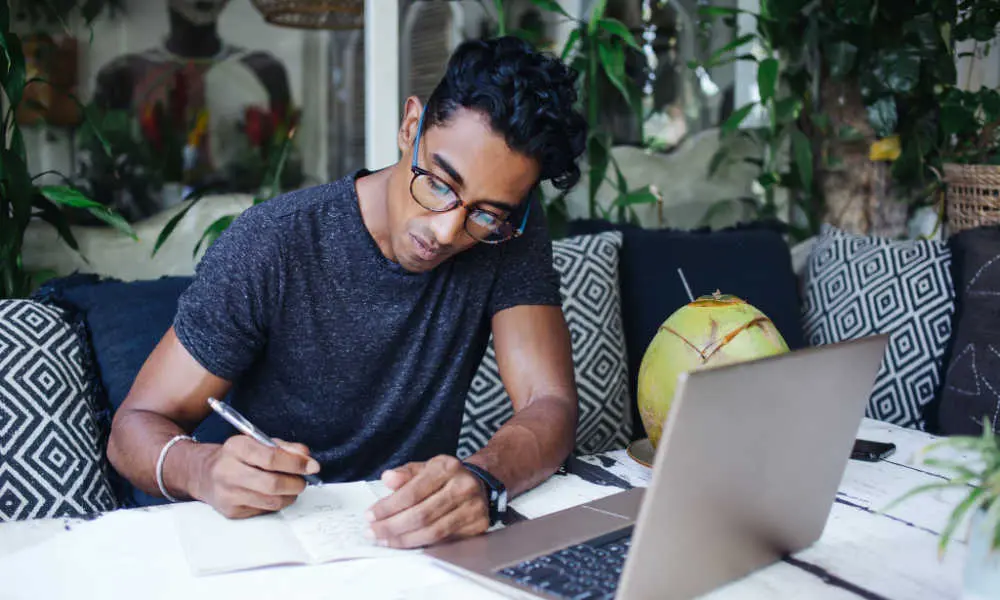A man wearing glasses writes in a notebook beside an open laptop.