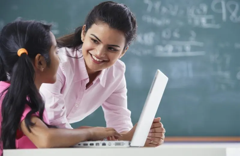 A teacher smiles at a student who is working on a laptop in a classroom.