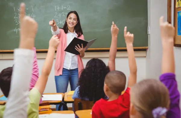 A teacher points to a classroom full of students with their hands raised.