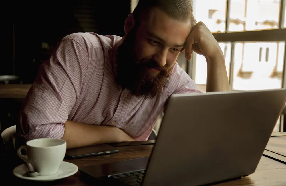 A man sits at a wooden table with a cup of coffee while he works on his laptop.