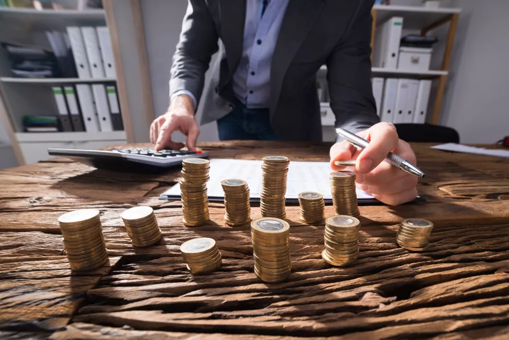 Person at a wooden desk calculating finances with stacks of coins and a calculator.