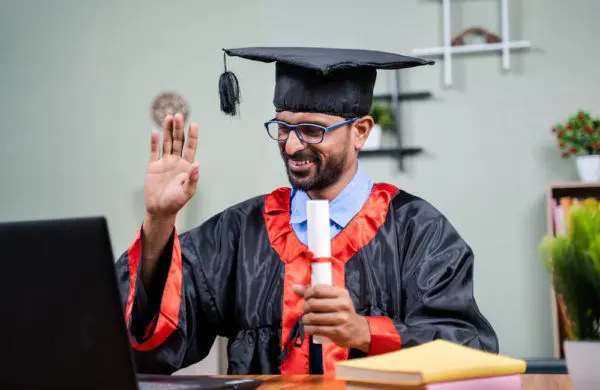 A man wearing a cap and gown and holding a diploma waves at a laptop screen.