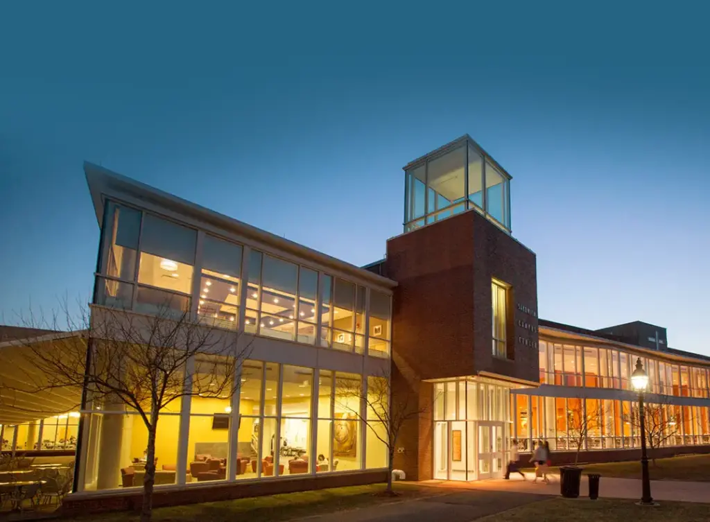 A Merrimack College building at dusk, with warm yellow light filling the building against a darkening blue sky.