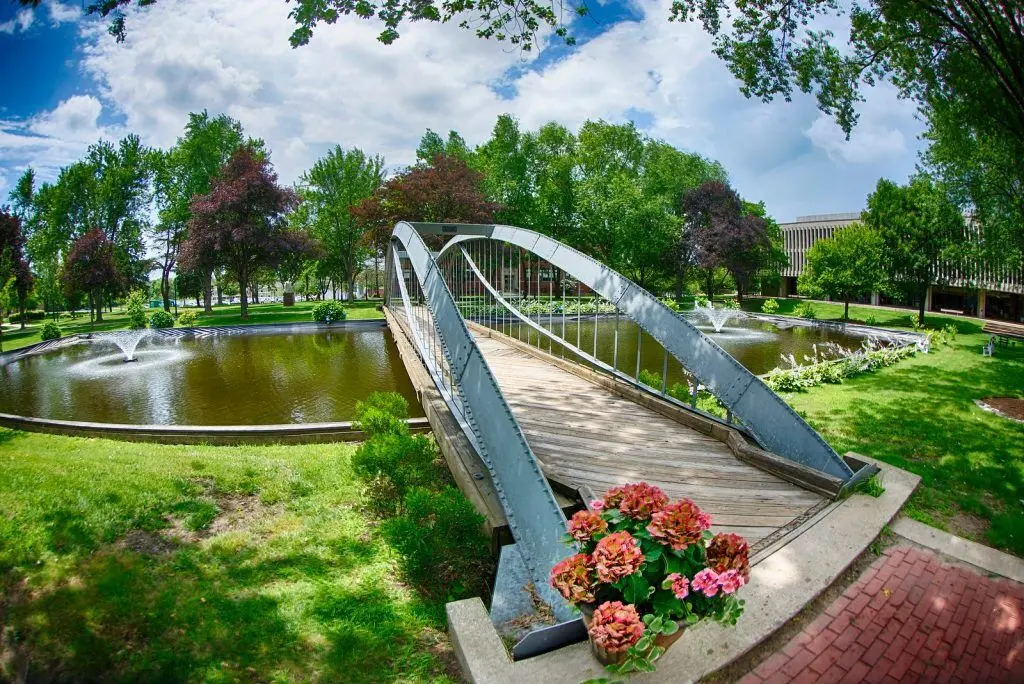 A bridge leads across a pond with fountains and flowers in the foreground on the Merrimack College campus.