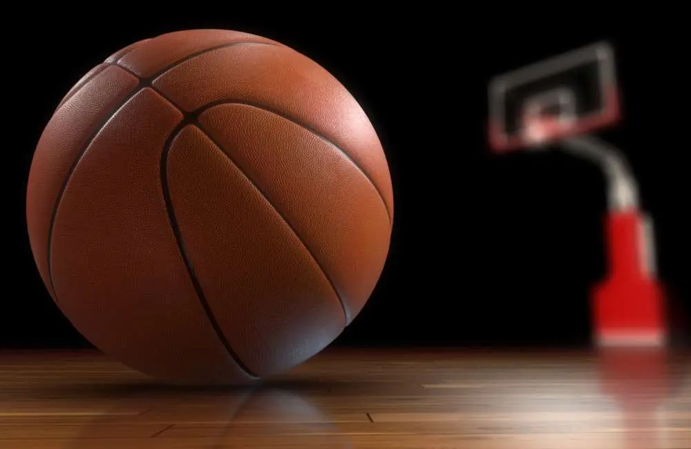 A closeup of a basketball on a wooden court floor with a basketball hoop in the background.