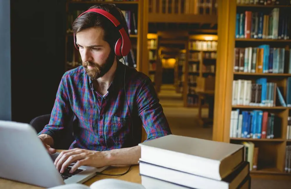 A man wearing headphones works at a laptop in a library.