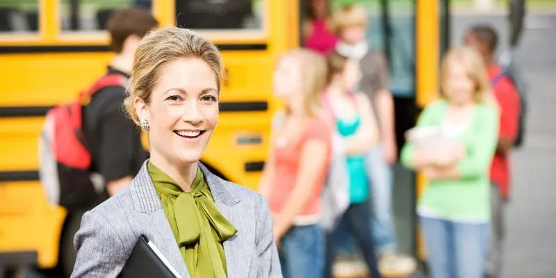 A principal smiles at the camera while students get off a yellow school bus in the background.