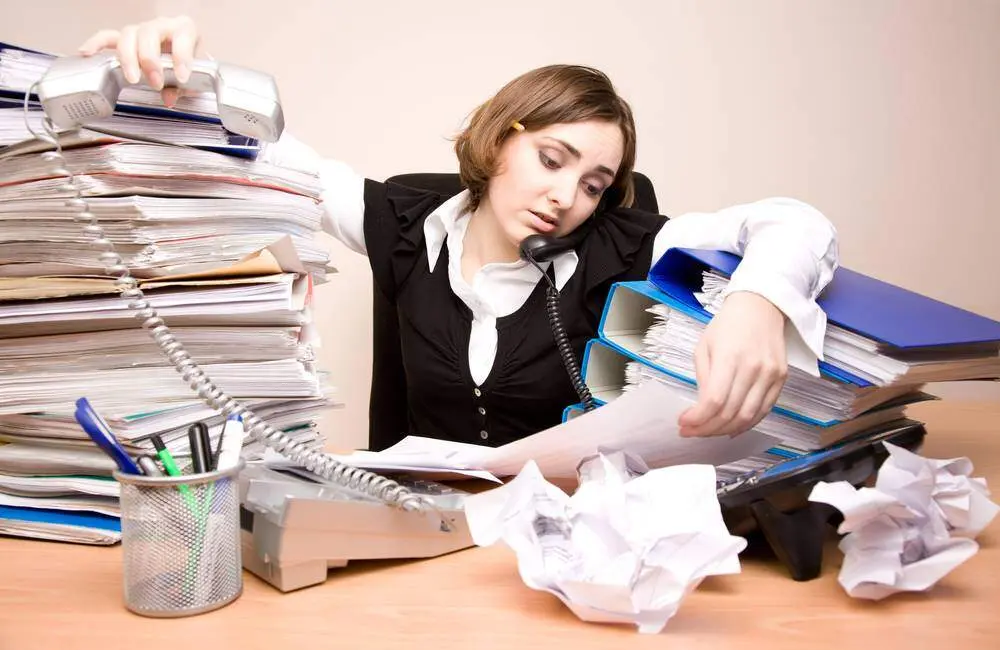 A woman speaks on the phone in the midst of stacks of papers, binders, an additional landline phone, and crumpled up pieces of paper.
