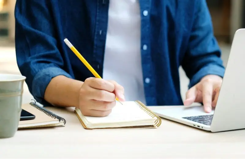 A closeup of one hand writing notes in a notebook while the other hand rests on the trackpad of a laptop.
