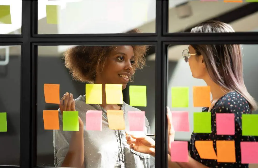 Two colleagues talk to each other in front of a wall of windows with different colored post-it notes stuck onto them.