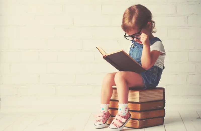 A young child wearing an adult's glasses sits on a stack of books with one open on her lap.