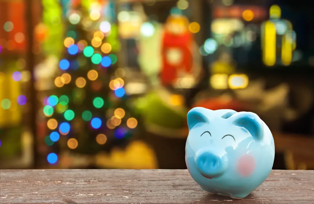 A blue piggy bank sits on a table with the glow of colored lights on a Christmas tree in the background.