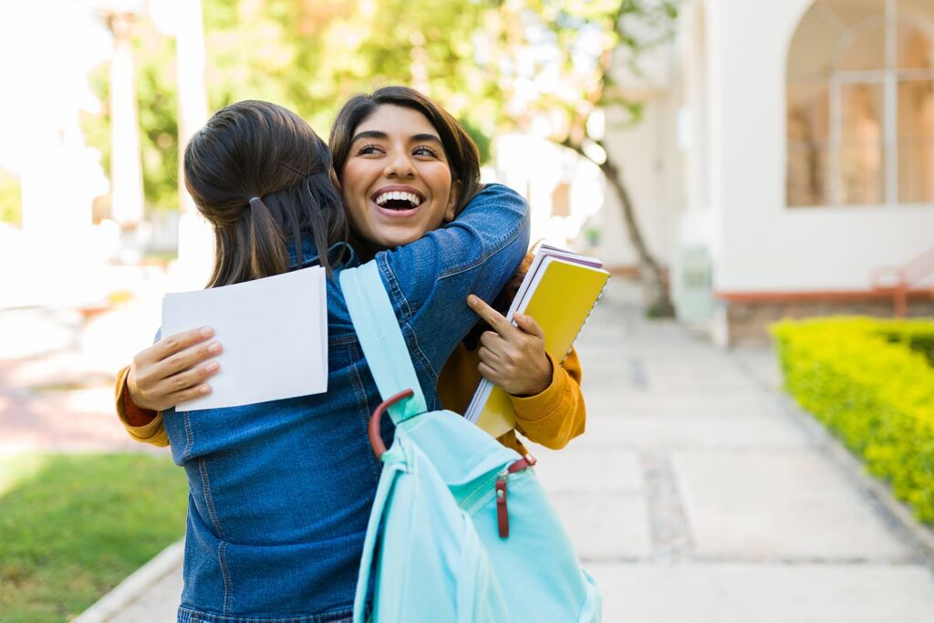 Two female students in a campus common space hug and smile.