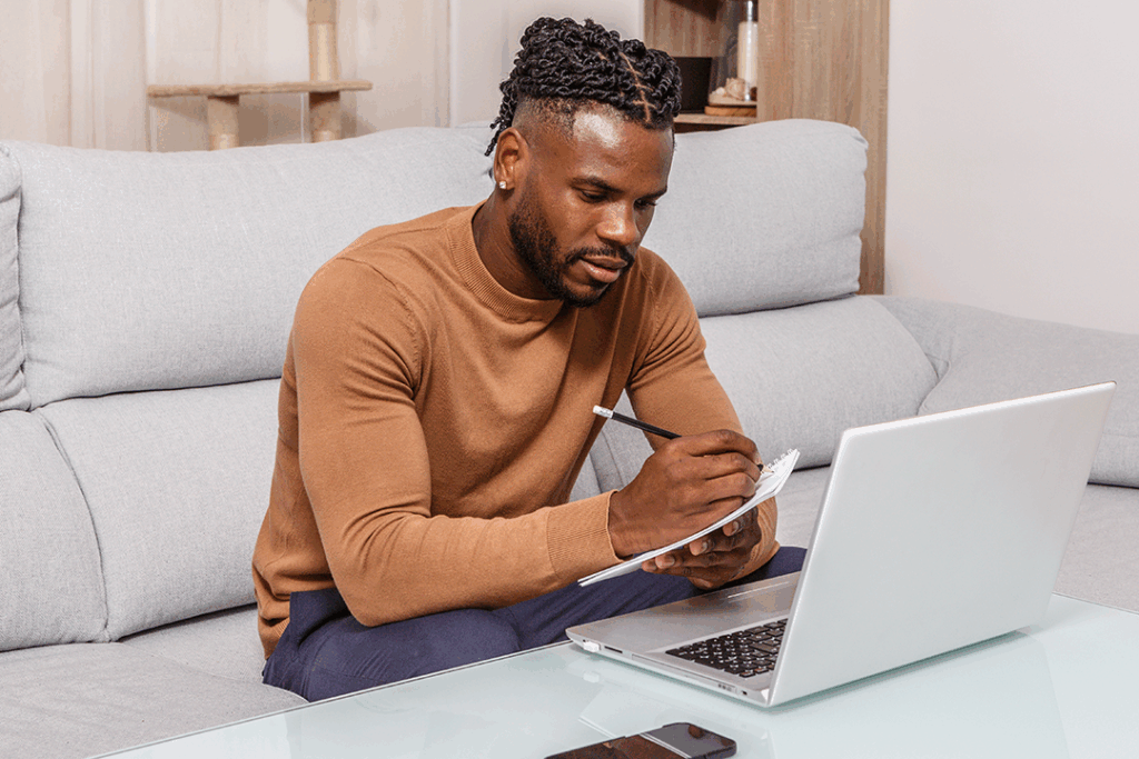Person sitting on a sofa, writing notes on paper while looking at an open laptop on the table in front of them.