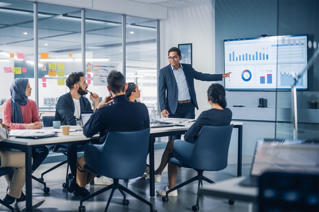 A man in a suit presents charts and graphs on a screen to a diverse team seated around a conference table in a modern office meeting room.