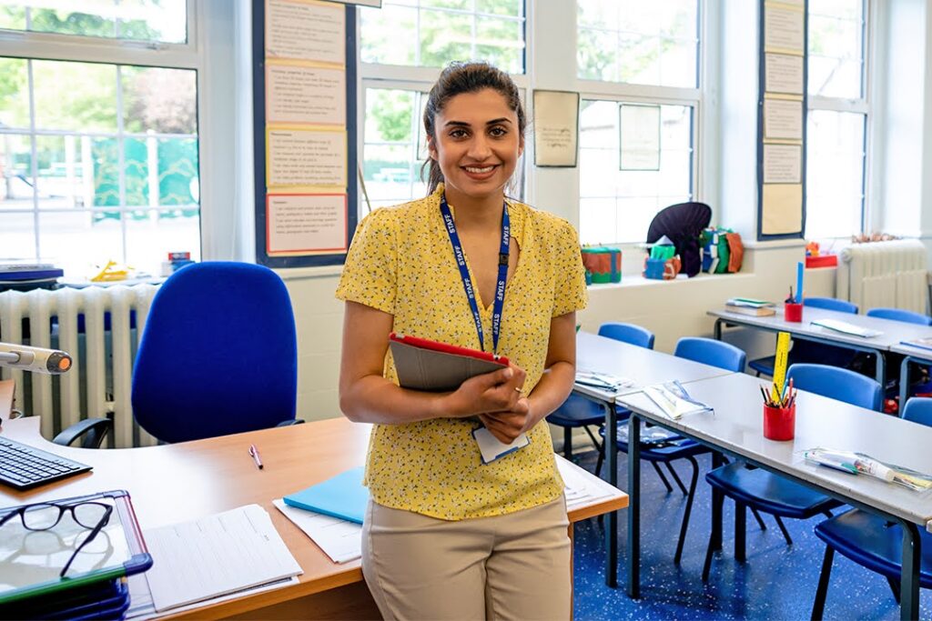 A smiling teacher holding a clipboard stands before her desk at the front of an empty classroom.