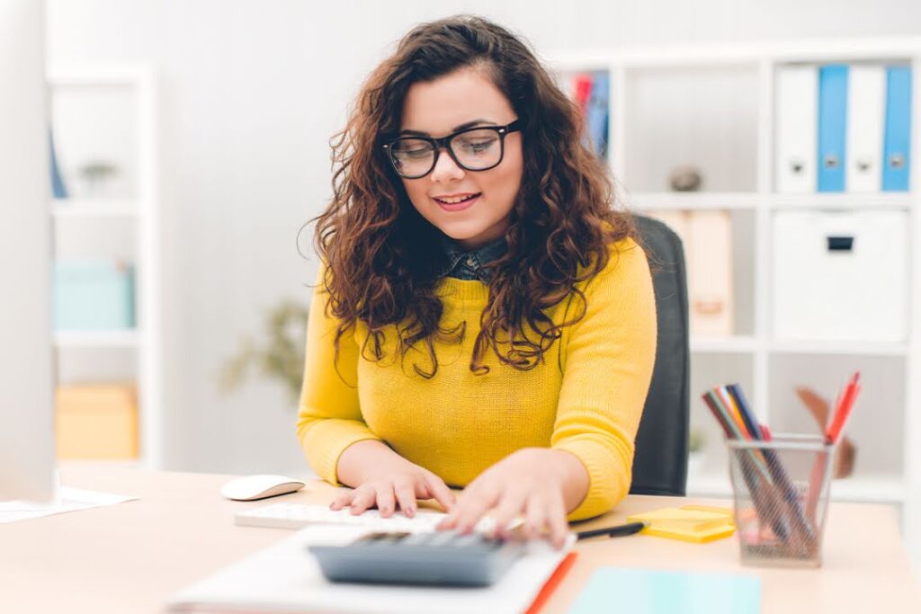 A smiling woman seated at a computer station in a modern office space uses a calculator.