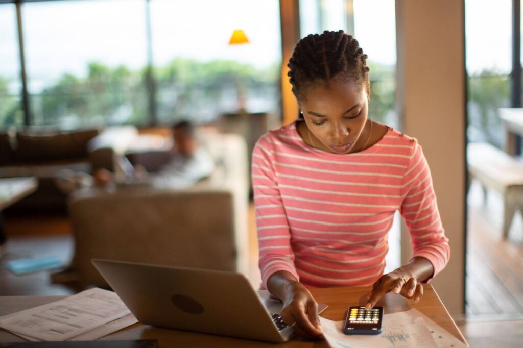 A woman seated at a table with a laptop in a cozy, sunlit room uses a calculator while looking at financial documents.