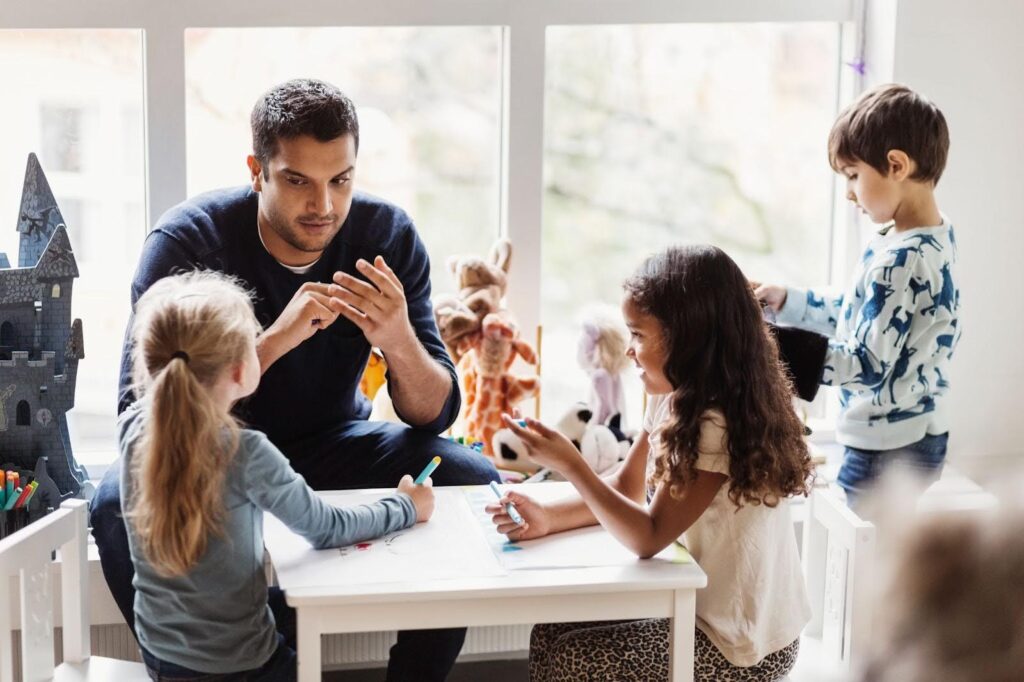 A man sits with young children at a small table, engaging in a learning activity with markers as another child plays nearby in a bright room with toys.